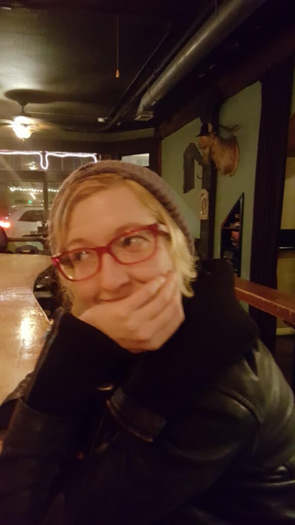 Woman in red glasses grins behind her hand, leaning at a bar in a leather coat.